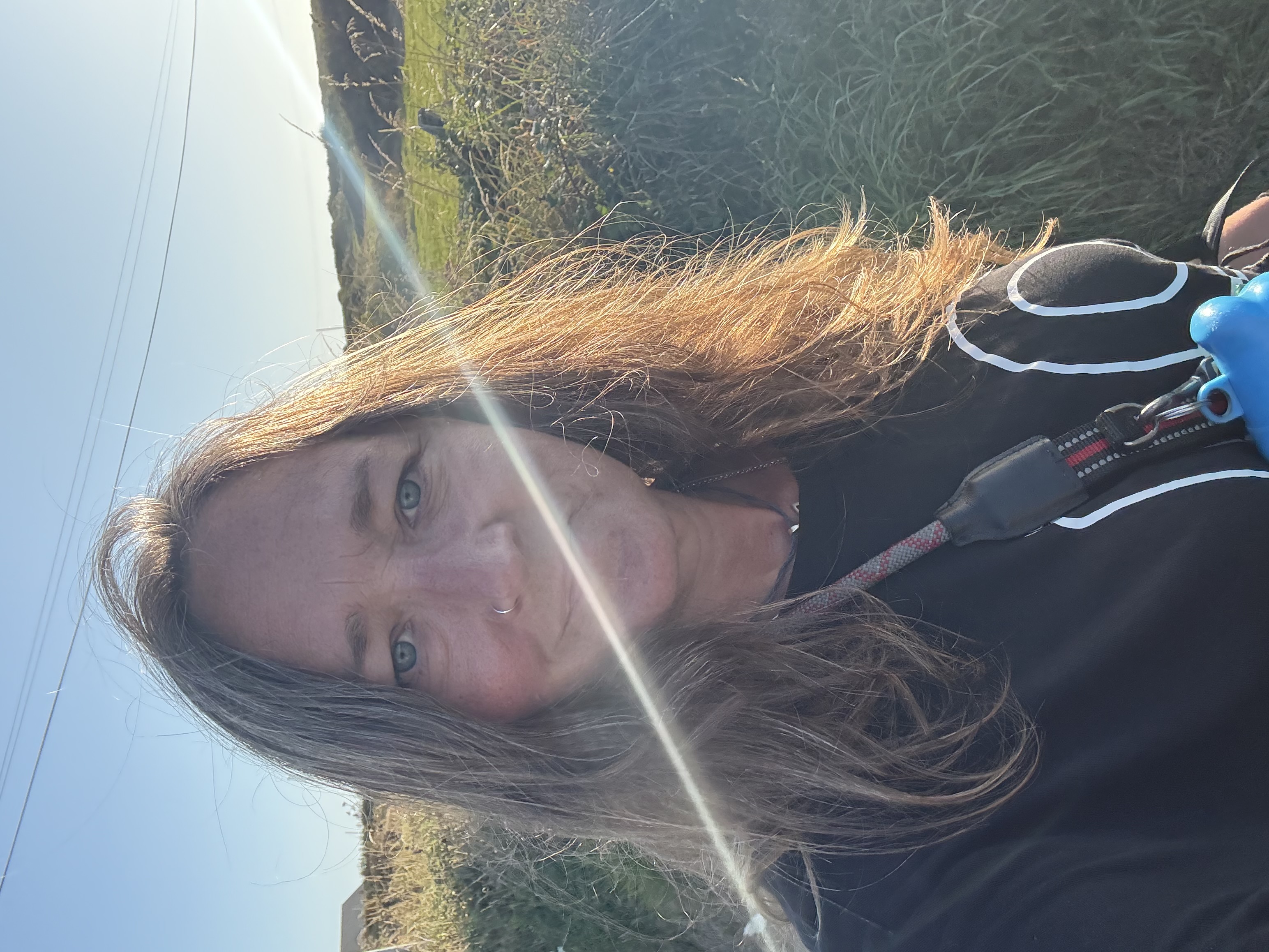 Ali Knight, a white woman with long blonde flecked hair and a nose ring, looking directly at the camera outdoors on a coastal path in golden evening light, holding a dog lead, with a lens flare across the image and wild grasses and blue sky behind her